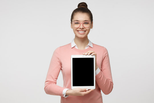 Studio picture of positive female isolated on grey background standing in casual clothes holding tablet and showing it blank screen with happy smile as if advising product or service