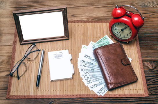 Empty photo frame with copy space, red alarm clock and wallet with money on wooden table background.