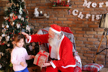 Gorgeous female child with great joy presents box Christmas surprise to Santa Claus who sits on...