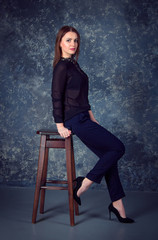 Portrait of beautiful young brunette woman, wearing black blouse and dark blue pants, sitting on a wooden chair in a room with gray wall 