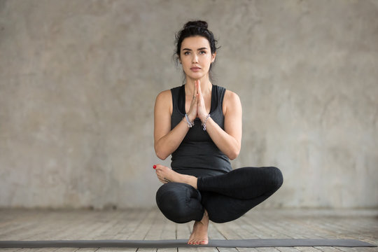 Young sporty woman practicing yoga, doing Half Lotus Toe Balance exercise, Toestand pose, working out, wearing sportswear, black pants and top, indoor full length, gray wall in yoga studio
