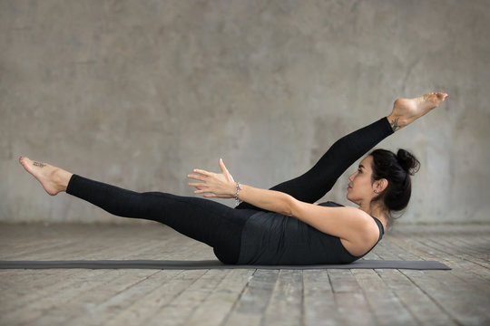 Young Sporty Woman Practicing Fitness, Doing Alternate Leg Stretch Exercise, Warm Up Pose, Working Out, Wearing Sportswear, Black Pants And Top, Indoor Full Length, Against Gray Wall In Sport Studio