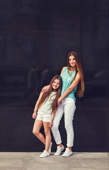 Young beautiful mom with her daughter wearing t-shirt and jeans posing against concrete wall, minimalist street fashion style, clothing for parent and child.