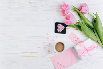 Pink tulips, envelope, gift, cup of coffee and paper hearts on white wooden background, copy space and flat lay. Mother's Day concept.