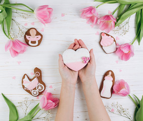 Female hands holding heart. A composition for newborns on a wooden white background with pink tulips, hearts and a cookies, copy space and flat lay. It's a girl.