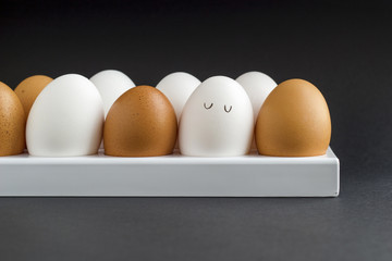 Fresh chicken egg in container on black paper background. Concept for Easter with copy space. White and brown eggs. Painted cute face on a white egg. Drawn cartoon eyes. Frontal view photo.