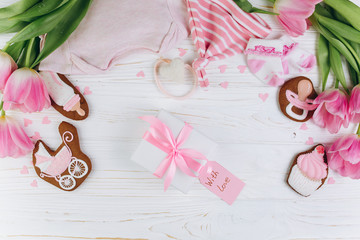 A composition for newborns on a wooden white background with gift, clothes, pink tulips, hearts and a cookies, copy space and flat lay. It's a girl.