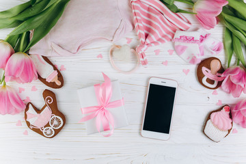 A composition for newborns on a wooden white background with gift, smartphone, clothes, pink tulips, hearts and a cookies, copy space and flat lay. It's a girl.