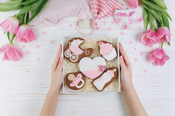 Female hands holding box with cookies. A composition for newborns on a wooden white background with clothes, pink tulips and hearts, copy space and flat lay. It's a girl.