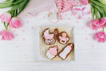 Female hands holding box with cookies. A composition for newborns on a wooden white background with clothes, pink tulips and hearts, copy space and flat lay. It's a girl.
