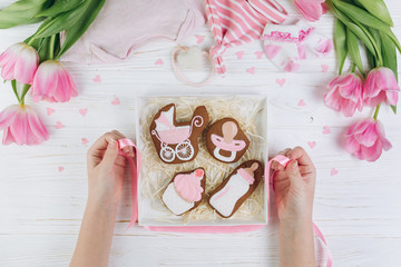 Female hands holding box with cookies. A composition for newborns on a wooden white background with clothes, pink tulips and hearts, copy space and flat lay. It's a girl.