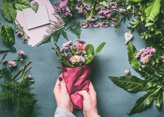 Female hand making beautiful bouquet of pink flowers on florist workspace, top view. Woman wrap bouquet in wrapping paper, step by step