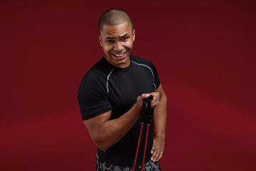 Isolated studio portrait of clean shaven young dark skinned male athlete with strong muscular shoulders working out using resistance elastic band, strengthening arm muscles, clenching teeth