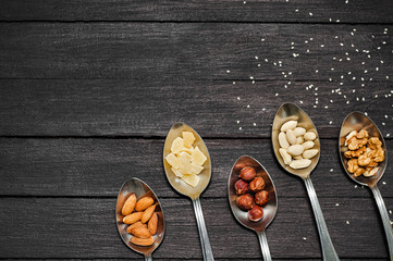Different kinds of nuts. On a dark wooden table lie spoons with nuts of almonds, walnuts, hazelnuts, dried pineapples, peanuts. The table is strewn with seeds of flax and sesame.