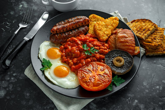 Full English breakfast with bacon, sausage, fried egg, baked beans, hash browns and mushrooms in black plate. cup coffee.