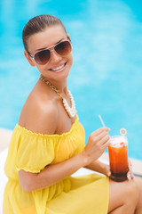 Beautiful tanned young woman with perfect body wearing in a short yellow sarafan and sunglasses, with a glass of fruit juice,relaxing near swimming pool with blue water. outdoor shot. copy space