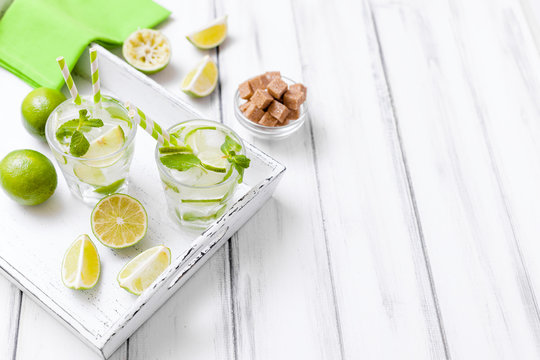Caipirinha, mojito cocktail with lime, brown sugar, ice and mint leaves in beautiful glasses, cut green citrus on white wooden background. Summer alcohol drink. Close up photography. Selective focus