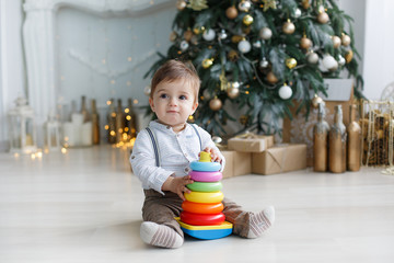 A little boy sits on a white glossy floor near green, Christmas tree with gift boxes under it, against the background of festive decorations and lights, one collects a multi-colored plastic pyramid