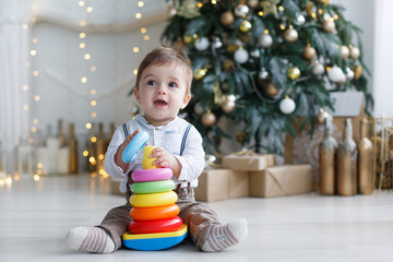 Fototapeta na wymiar A little boy sits on a white glossy floor near green, Christmas tree with gift boxes under it, against the background of festive decorations and lights, one collects a multi-colored plastic pyramid