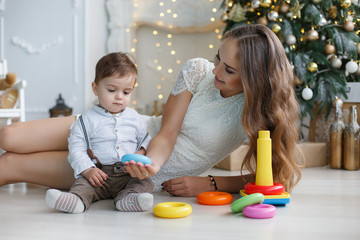 Naklejka premium Mother playing with her son on the eve of the New Year. Mother teaches young son to collect the multicolored pyramid. Happy family on Christmas tree background.