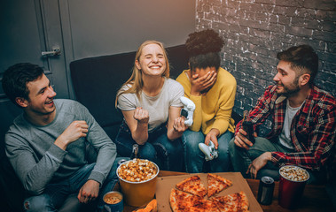 An emotional picture wher blonde girl wins and afroamerican girl loses. One of them is happy about winning the game while the other one is upset. Boys are just having some fun. Gamers. Night party.