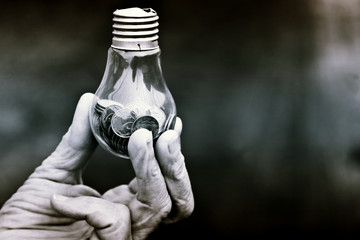 Black and white photo of a light bulb with coins in old wrinkled hands on a dark background