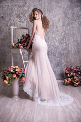 Beautiful bride in lace dress. Standing on the ladder. Floor-length dress. From the back. Open back. Bouquets of flowers. Gray background.