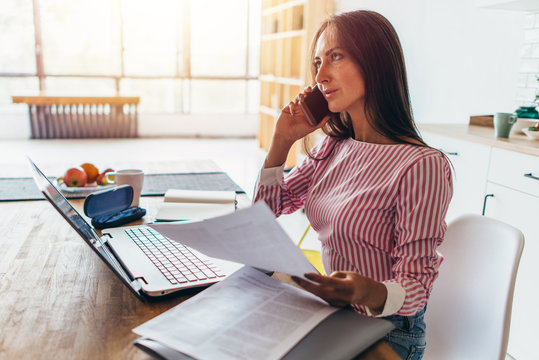 Thoughtful Woman Talking On Mobile Phone, While Working With Laptop From Her Home Office