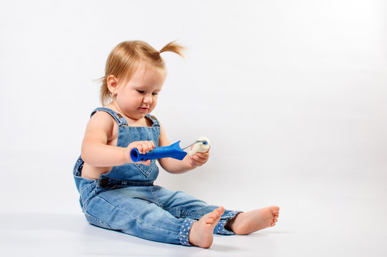 Little, cute baby girl dressed in denim overalls playing with paint roller