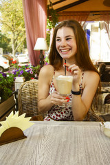 Beautiful young girl sits on the terrace of a cafe and drinks latte. She is happy and smiling. Sunny day. Flower decoration. French coffee house