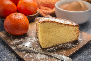 orange sponge cake with icing sugar on wooden plate on top of a table with tangerines and eggs