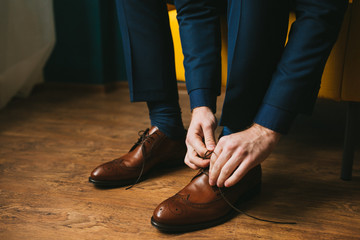 A man or a groom in a blue suit ties up shoelaces on brown leather shoes brogues on a wooden...