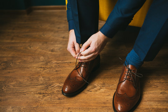 A man or a groom in a blue suit ties up shoelaces on brown leather shoes brogues on a wooden parquet background