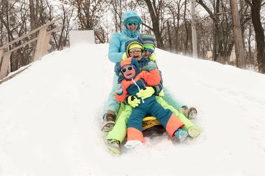 Happy mother with children in sunglasses and bright blue ski suit. Mother and boys are happy together. Kids ride a yellow sled in the forest with their mom. Guys are very happy to play outdoors
