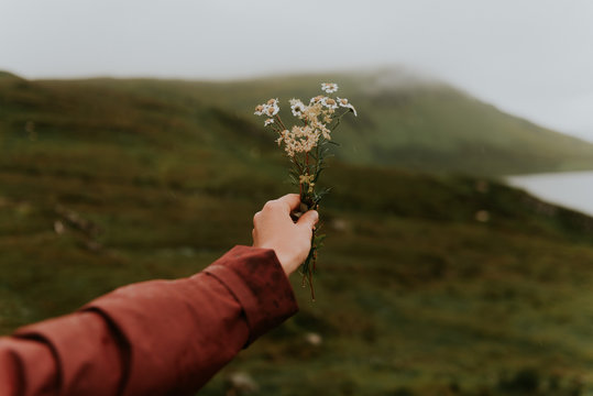 Woman wearing a red rain jacket holding a flower bouquet in front of a green field bokeh unsharp background in Scotland, UK
