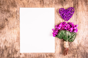 Obraz premium The first spring flowers and wicker heart on a wooden board. Flowers and blank sheet of paper. Romantic concept.