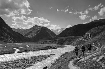 Black and white photo of group of backpackers walking along the trail on the rivershore. Sun is shining brightly and high mountain ridge could be seen on the beckground.