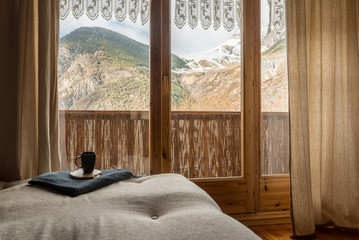 Vista de una habitación con un gran ventanal con vistas a unas impresionantes montañas cubiertas de nieve y sobre un sofá una taza negra de café. 