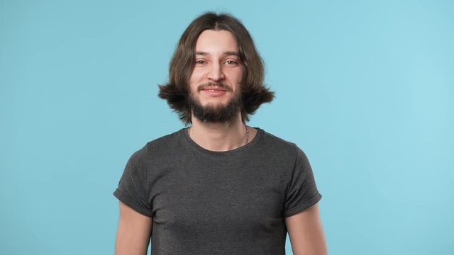Portrait of content bearded guy in casual grinning and greeting with waving hand on camera, over blue background. Concept of emotions