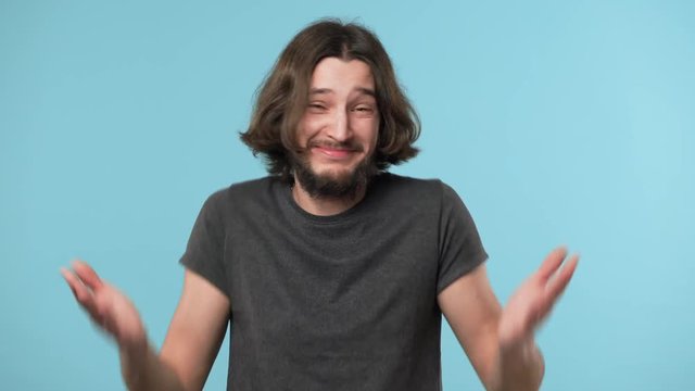 Portrait of innocent guy with beard and brown hair shrugging and gesturing with both hands meaning oops sorry have no idea, over blue background. Concept of emotions