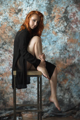 The beautiful red-haired woman sit on a high chair baring the leg. The production photo with studio light