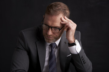 Nervous, overwhelmed businessman portrait.  Stressed businessman hand is on his forehead while sitting at dark background and thinking very hard. Professional man wearing suit.