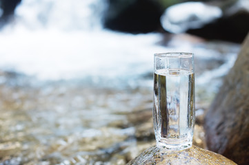 A transparent glass glass with mineral mountain river water stands on a stone beside the mountain river creek