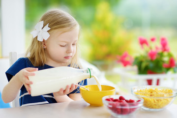 Cute little girl enjoying her breakfast at home. Pretty child eating corn flakes and raspberries and drinking milk before school.