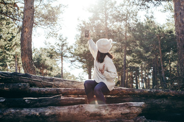 Young and pretty woman using her mobile phone outdoors, having fun taking herself pictures on a winter day with seated in a wooden trunk. Lifestyle.