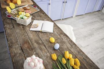 Kitchen bar table with opened notebook, flowers, glazed cookies and decorated basket with painted eggs. Copy space in right side