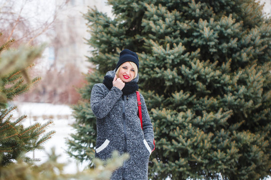 A hipster girl in a black hat in a gray coat looks at the camera and smiles outdoors. Portrait of a beautiful modern girl with red lips in winter.