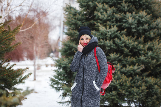 A hipster girl in a black hat in a gray coat looks at the camera and smiles outdoors. Portrait of a beautiful modern girl with red lips in winter.