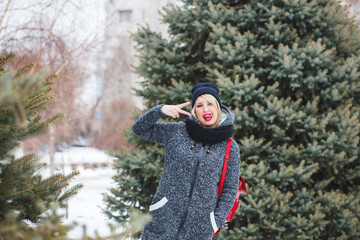 A hipster girl in a black hat in a gray coat looks at the camera and shows a gesture of fingers at the face outdoors. Portrait of a beautiful modern girl with red lips in winter.