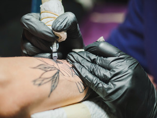 Tattoo artist at work. Woman in black latex glove tattooing a young man's hand with colorful...